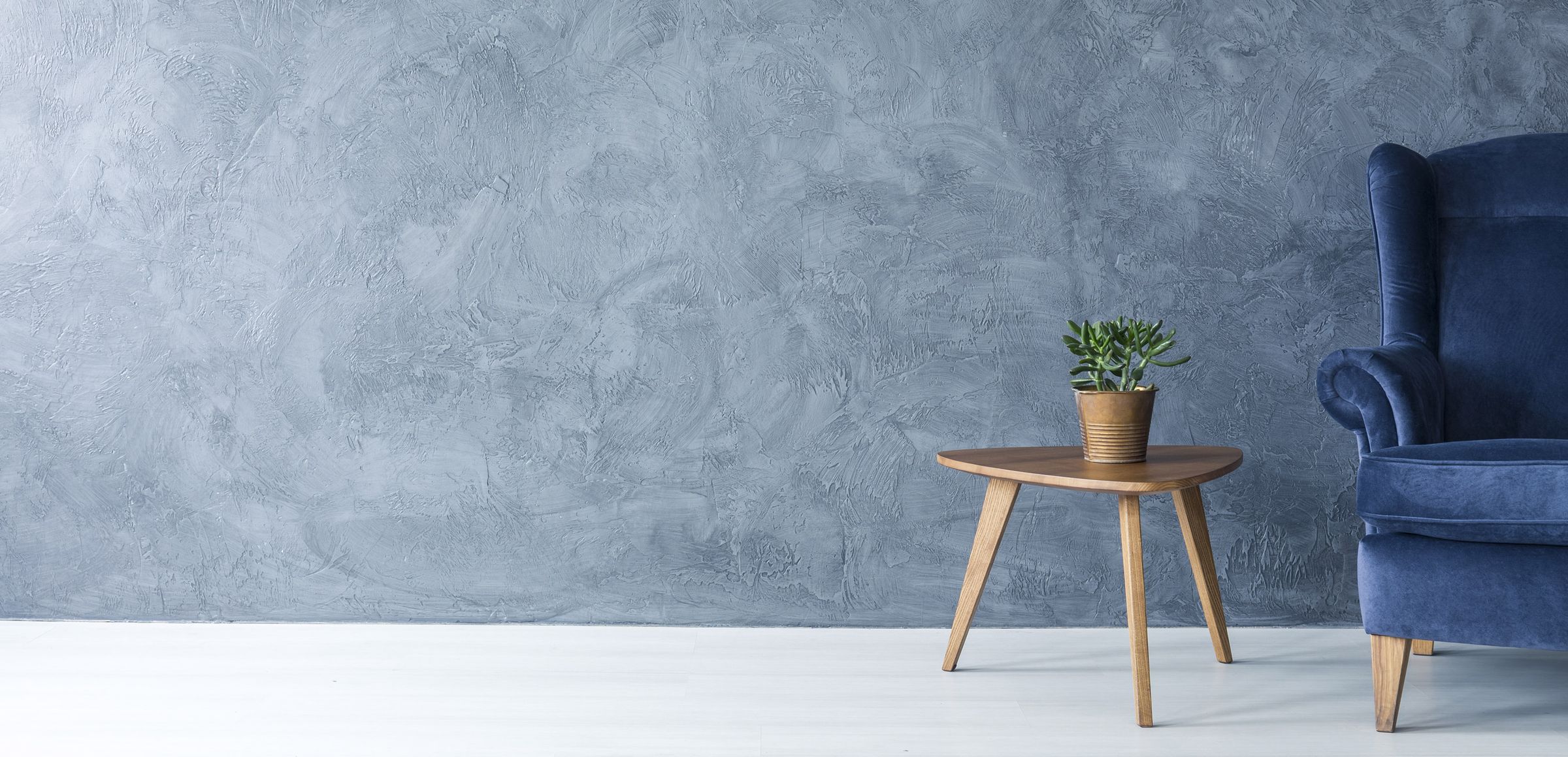 Ein blauer Sessel steht neben einem runden Holztisch mit einer Pflanze in einem Topf. Die Wand im Hintergrund ist grau und strukturiert.